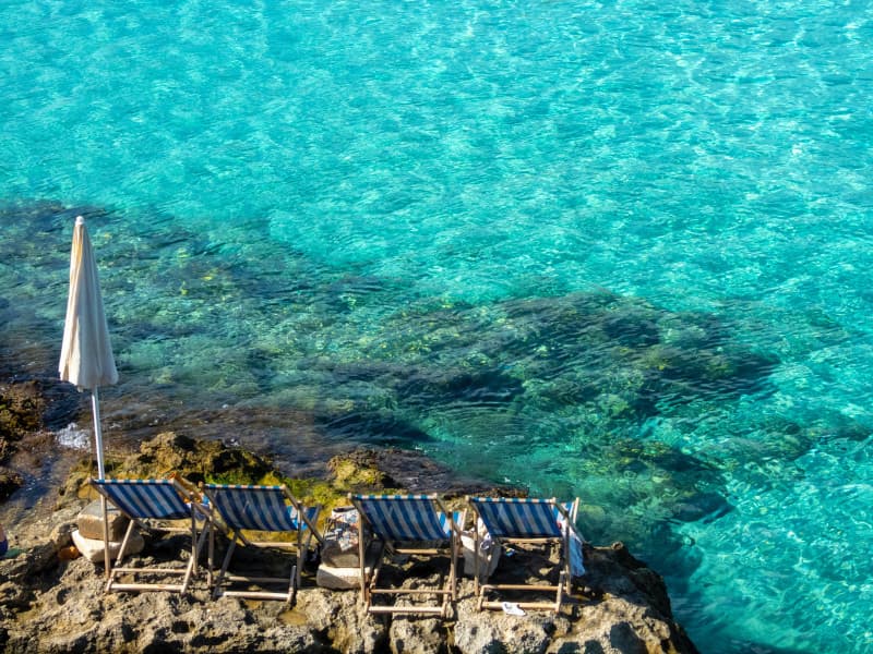 Photograph of a blue shallow sea with four deckchairs and an umbrella in the foreground on the rocks.